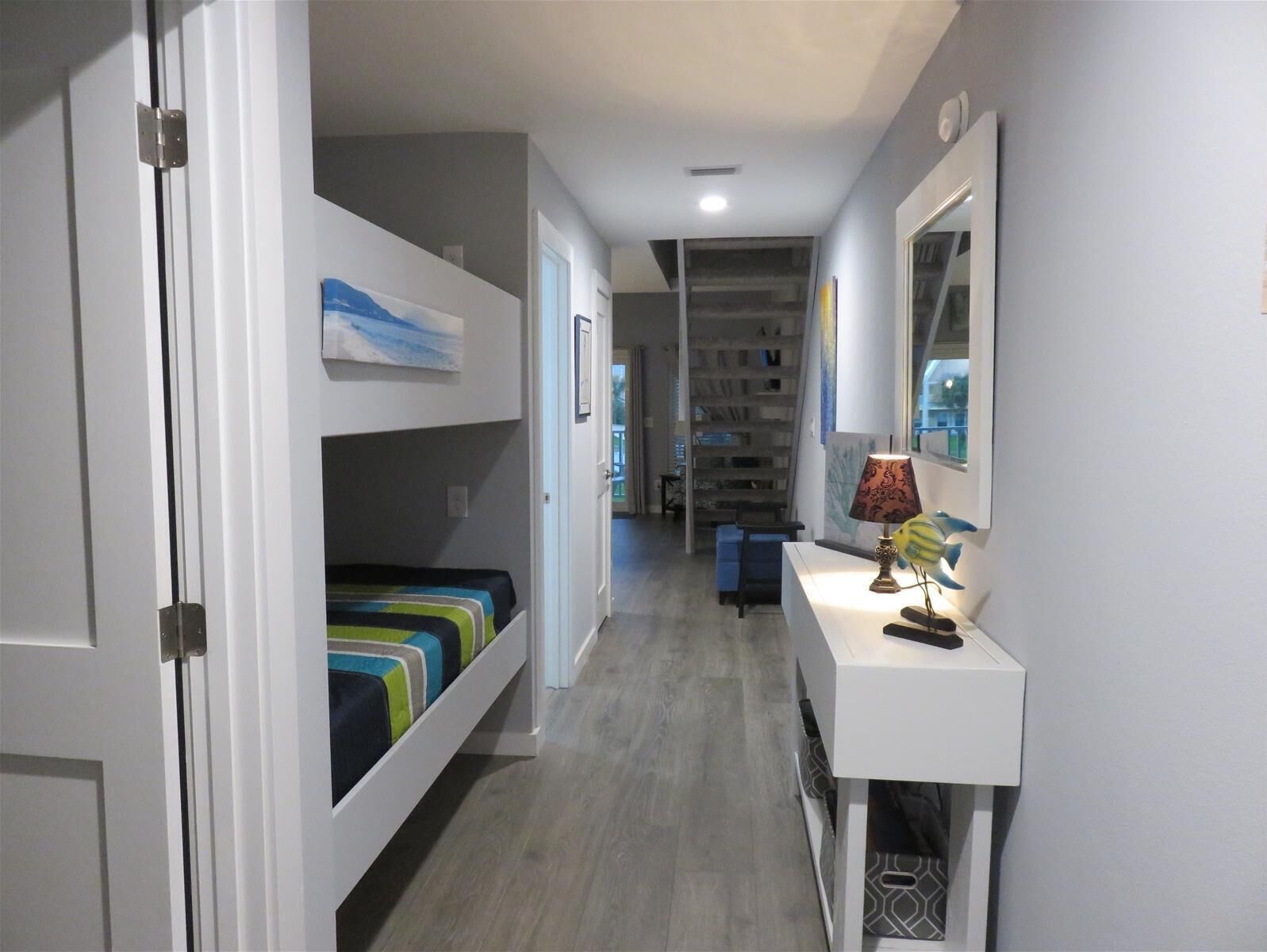 A narrow hallway with a bunk bed on the left, a desk with a lamp on the right, and a bookshelf in the background.