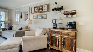 A living room with a beige sofa, a coffee station with a Keurig coffee maker, and a 'BEACH' sign on the wall. - Settle on the Sand (Miramar Beach)