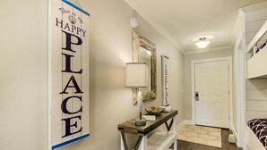 A hallway with a 'HAPPY PLACE' sign, a mirror, a lamp, and a door leading to a bedroom. - Settle on the Sand (Miramar Beach)