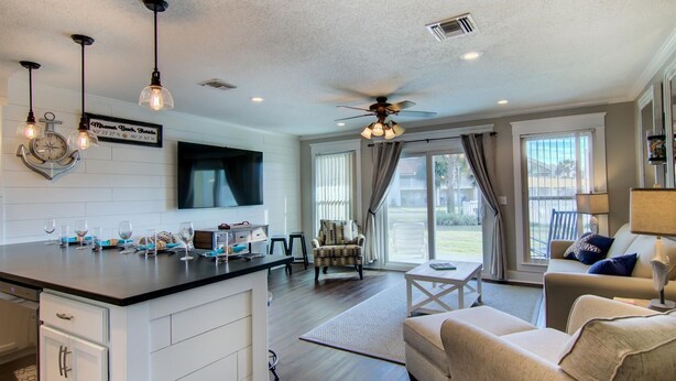 A modern kitchen with a central island, pendant lights, and a ceiling fan. There is a dining area with a sofa and a TV mounted on the wall. - Settle on the Sand (Miramar Beach)