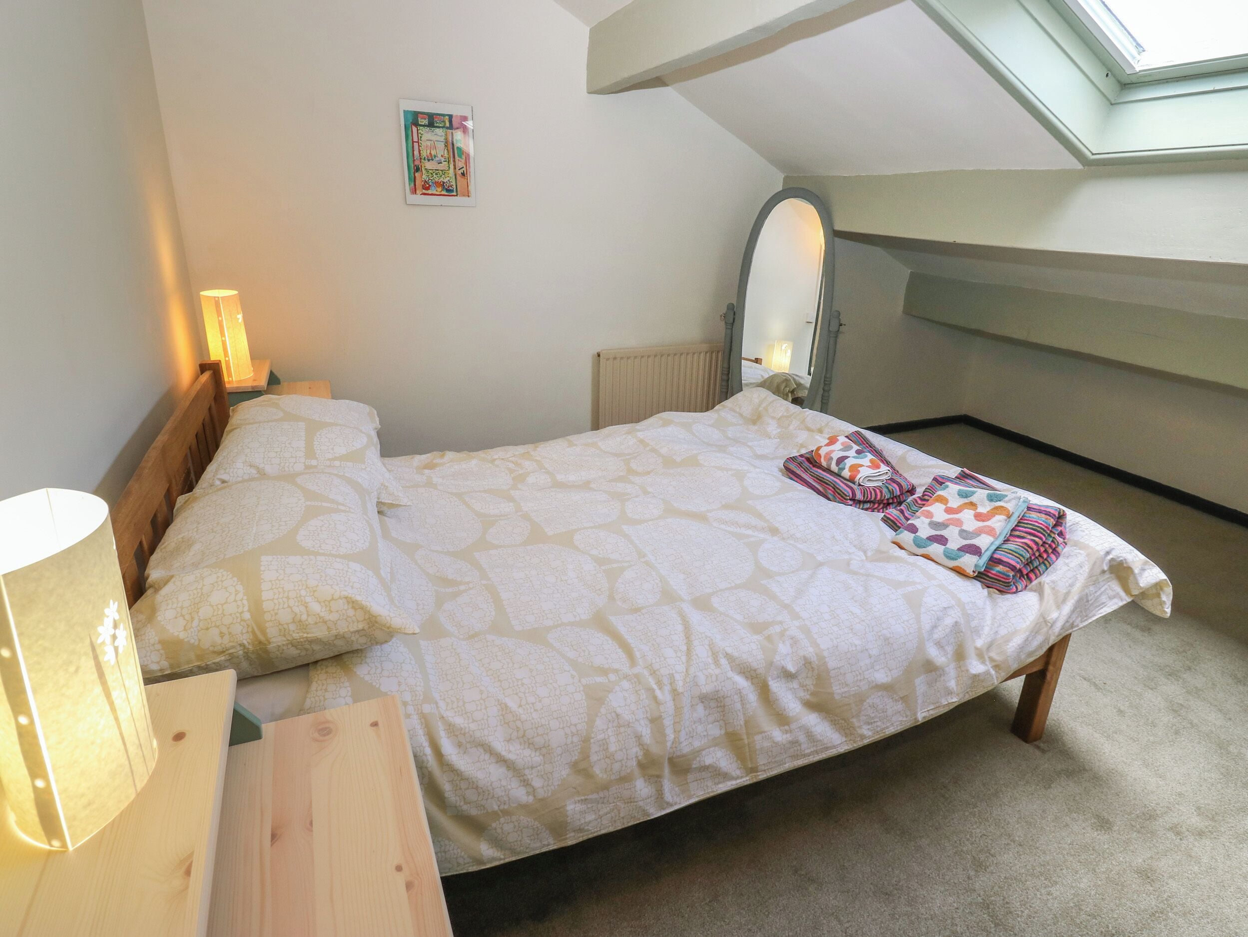 Un dormitorio con cama, lámpara de noche, espejo, radiador y tragaluz.