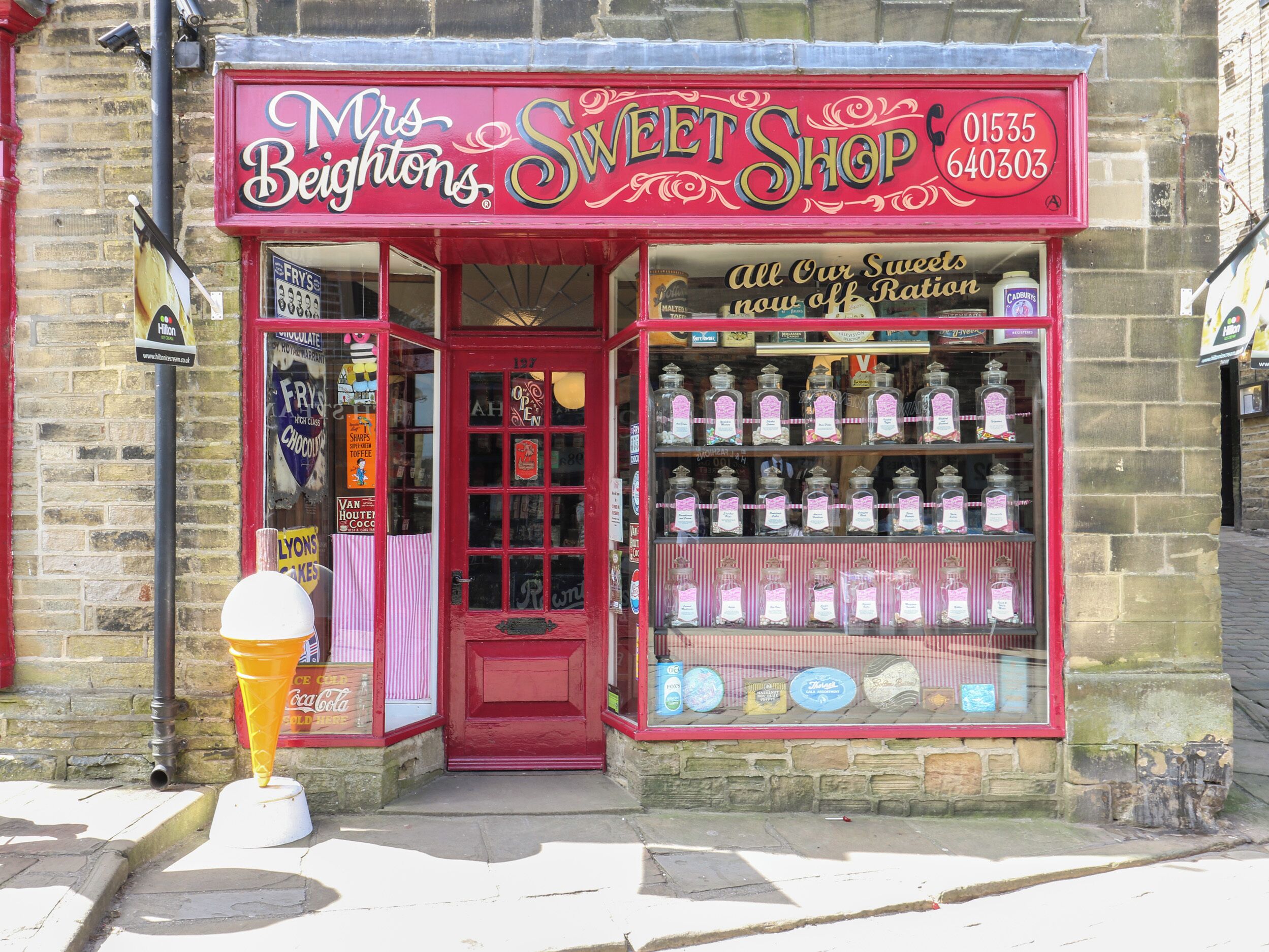 A storefront with a red sign reading 'Mrs Beightons Sweet Shop' and a display of sweets and cakes inside.