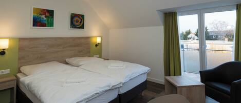 A hotel room with a double bed, two wall-mounted lamps, a small table, a chair, and a balcony with a view.