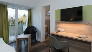 Quarto de hotel moderno com uma escrivaninha de madeira, uma cadeira preta, uma TV montada num painel verde e vista para o exterior através de uma porta deslizante.