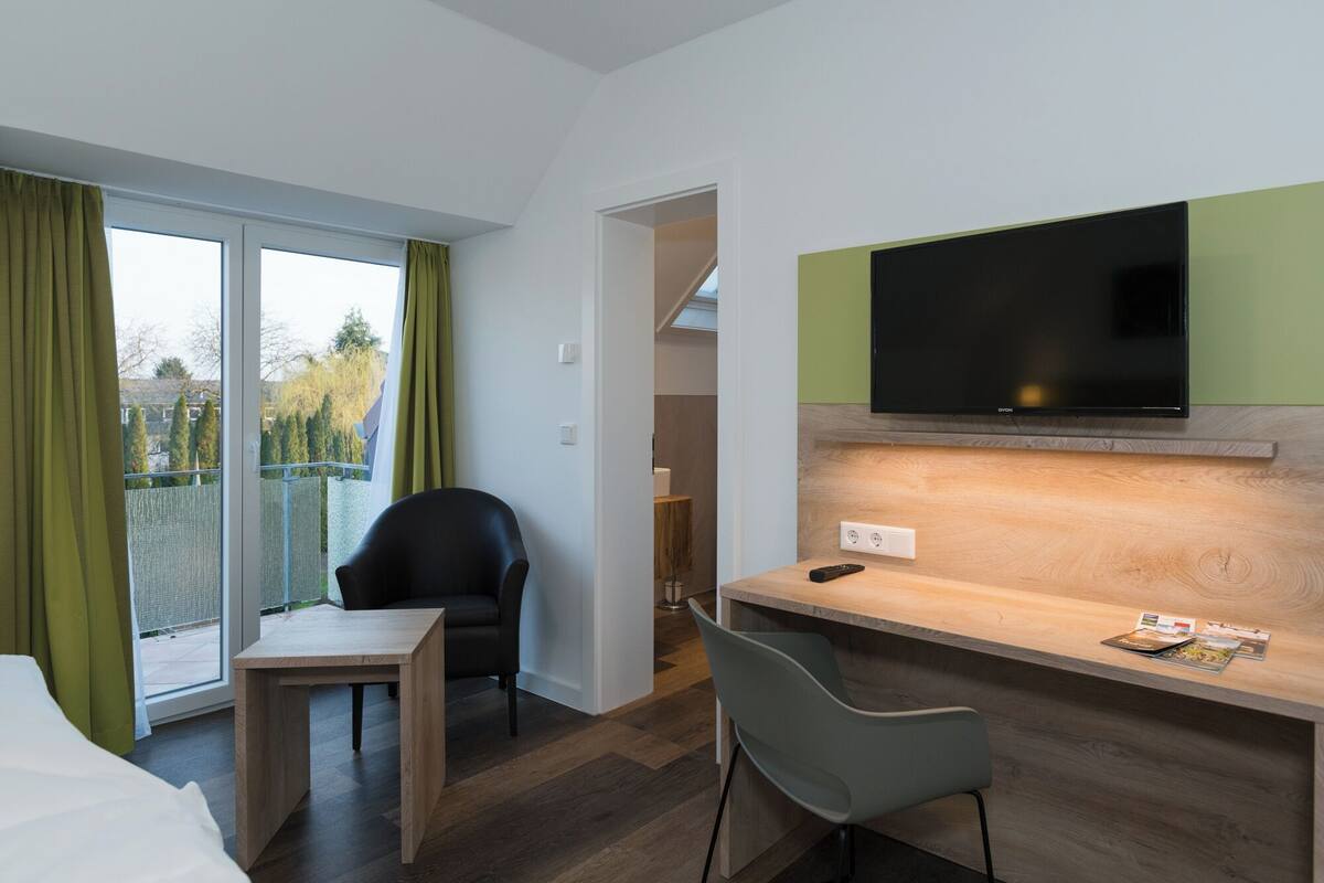 A modern hotel room with a wooden desk, a black chair, a TV mounted on a green panel, and a view of the outside through a sliding door.