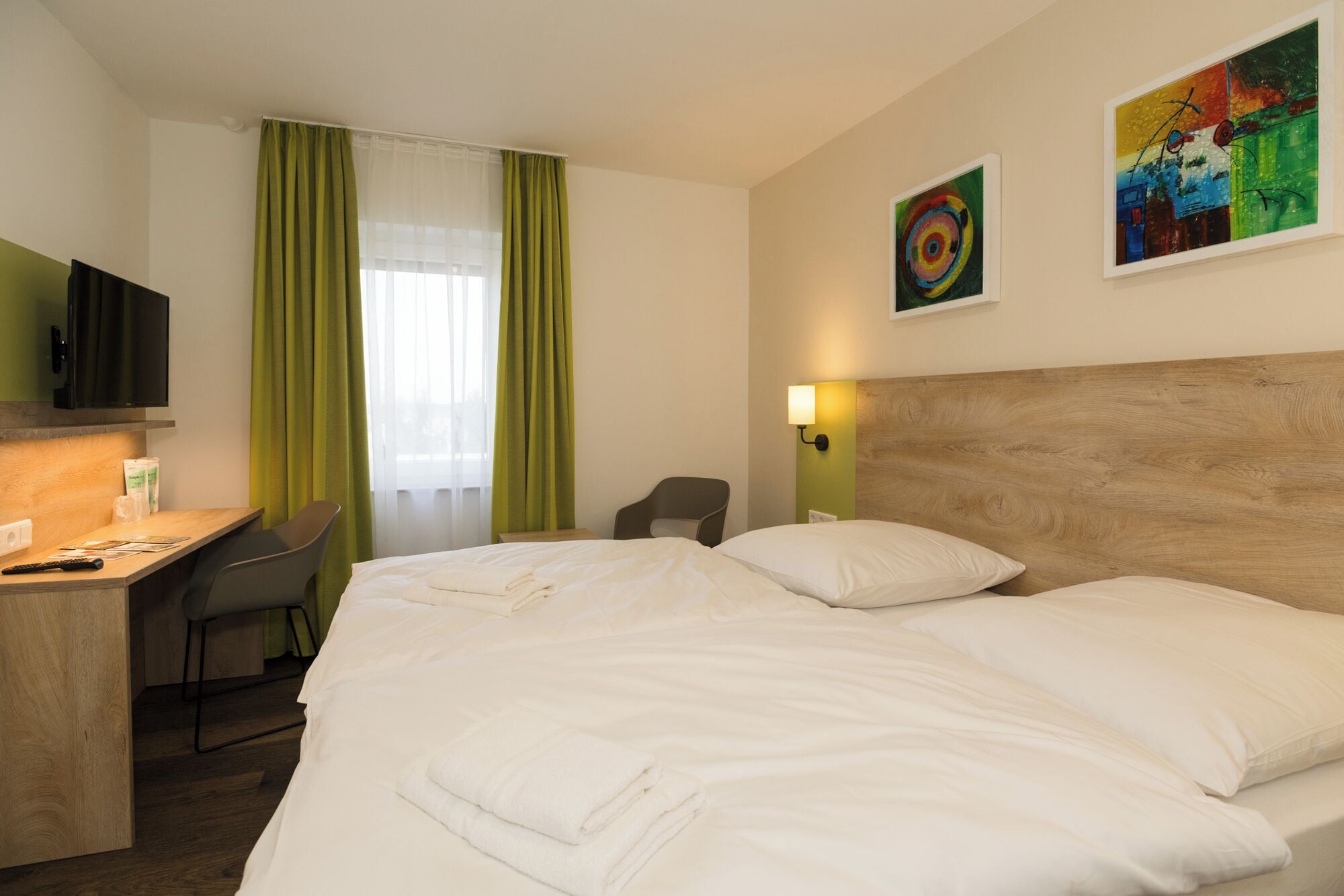 Une chambre d’hôtel équipée d’un lit, d’un bureau, d’une chaise, d’une télévision et de deux tableaux accrochés au mur.