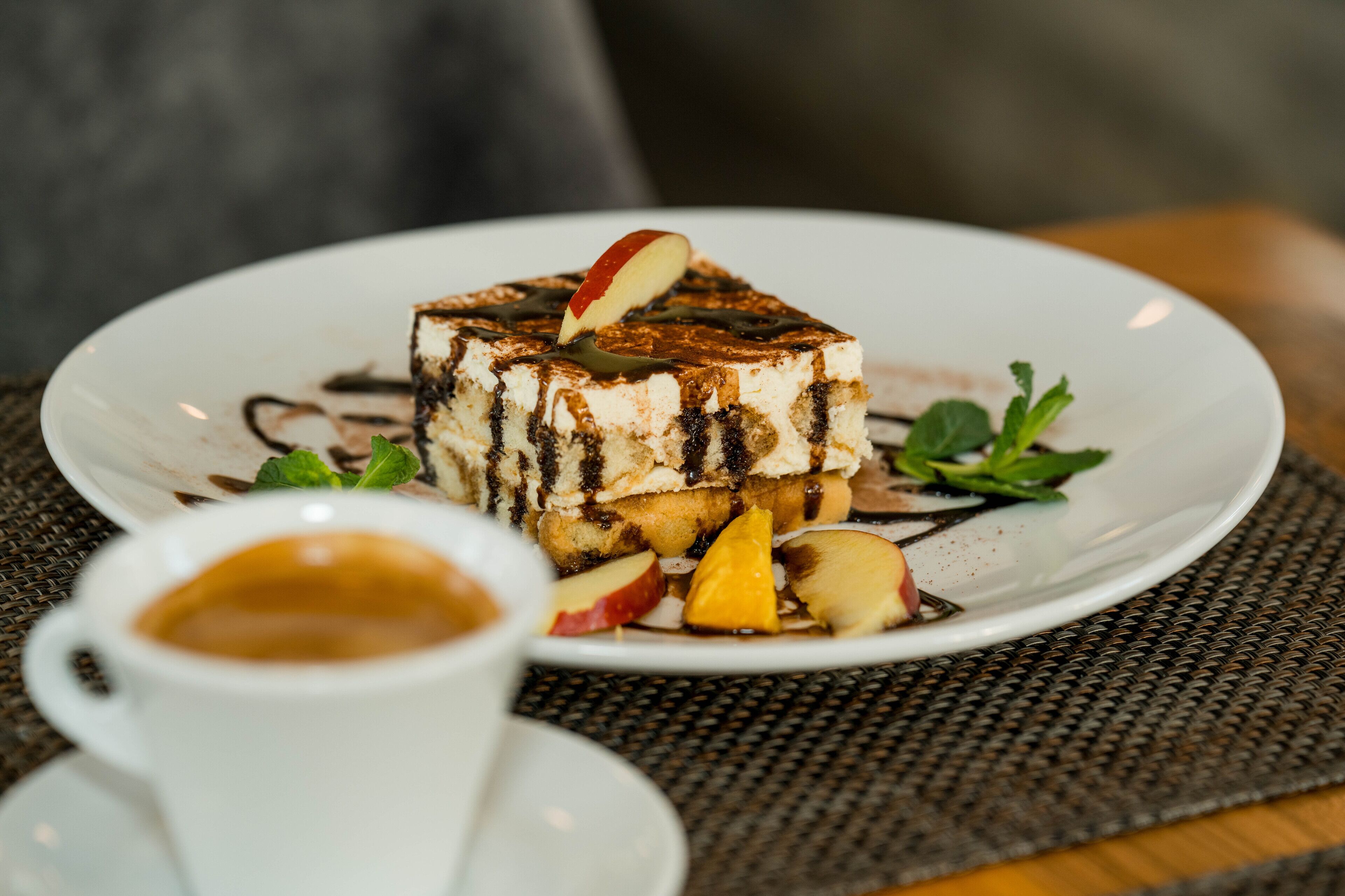 A dessert plate with a slice of cake, fruit, and chocolate drizzle on a white plate, accompanied by a cup of coffee.