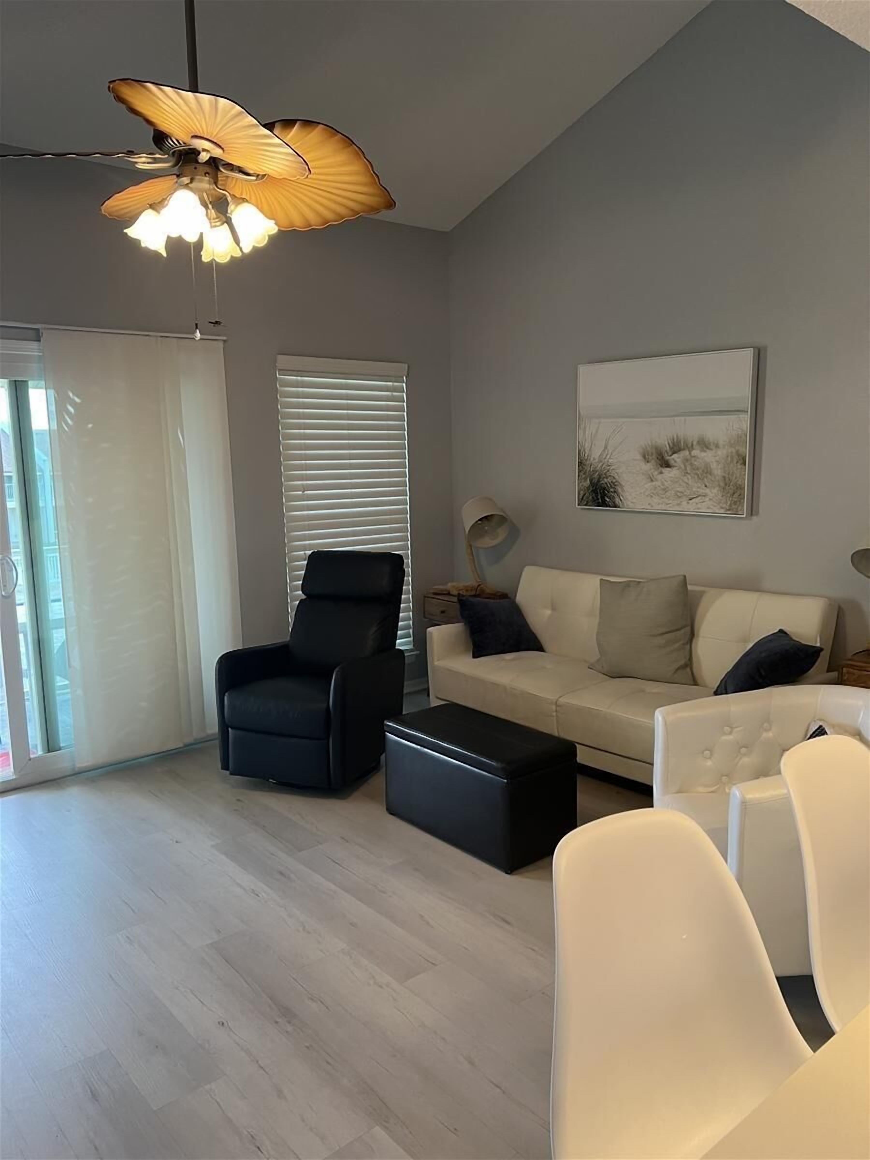 A modern living room with a black leather armchair, a beige sofa, and a white armchair. There is a black ottoman, a floor lamp, and a ceiling fan with a shell-shaped light fixture. A painting of a beach scene hangs on the wall.