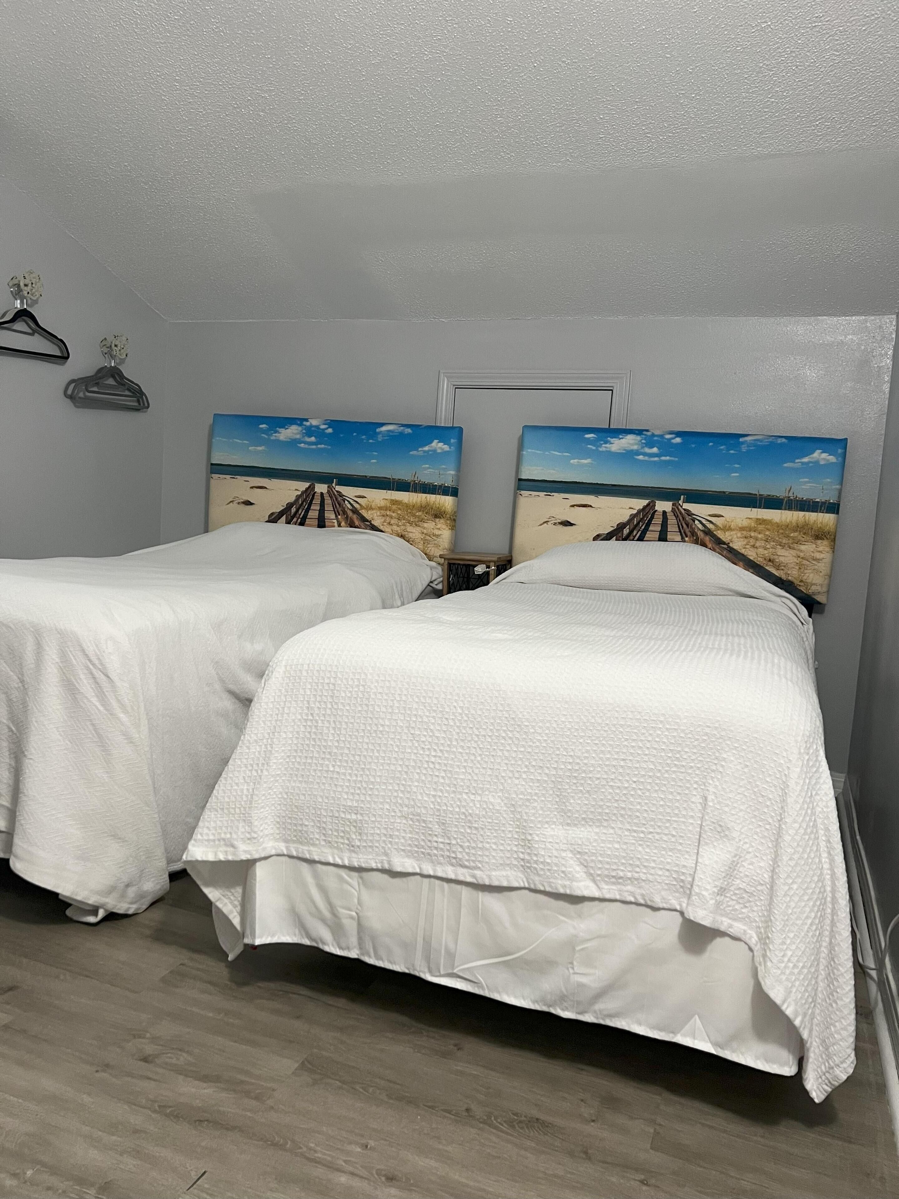Une chambre avec deux lits, chacun recouvert d’une courtepointe blanche, le tout adossé à un mur gris. Au-dessus des lits, on peut voir deux tableaux représentant une scène de plage.