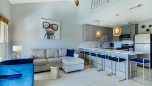 Een moderne woonkamer met een bank, een salontafel en een blauwe stoel. Er is een keukenhoek met een bar en een magnetron.
