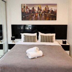 A hotel room with a large bed, two bedside tables, a cityscape painting, and a window.