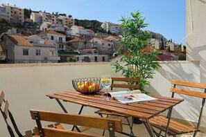 Une table en bois sur laquelle se trouvent un saladier de fruits et un verre de vin blanc, un pot de fleurs et un magazine, le tout placé sur un balcon donnant sur un coteau parsemé de bâtiments.