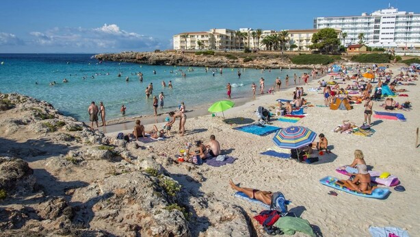 En strand med folk, der solbader og svømmer, et hotel i baggrunden og klippefyldt terræn.