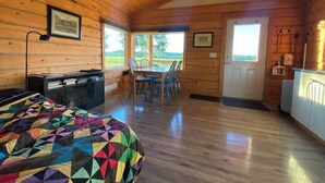L’intérieur chaleureux d’un chalet avec un plafond en bois, un coin repas comprenant une table et des chaises, un lit recouvert d’une couverture colorée et une vue sur l’extérieur grâce à de grandes fenêtres.