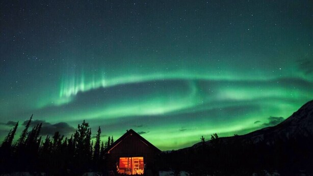 Une cabane dotée d’une fenêtre et d’une porte bien visibles, entourée de neige et d’arbres, sous un ciel nocturne illuminé par les aurores boréales.