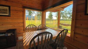 Une cabane en bois avec une table et des chaises, de grandes fenêtres donnant sur un champ verdoyant et des arbres.