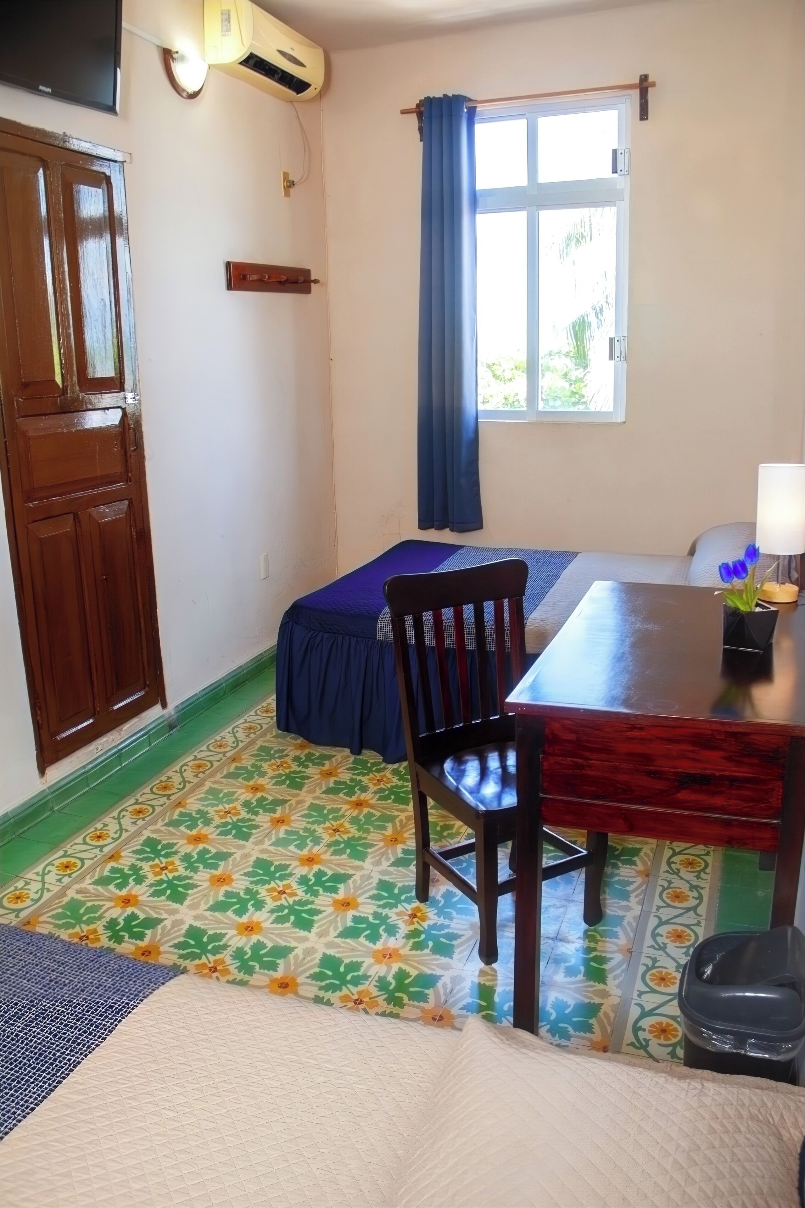 A room with a bed, a table, a chair, a window with curtains, an air conditioner, and a door.