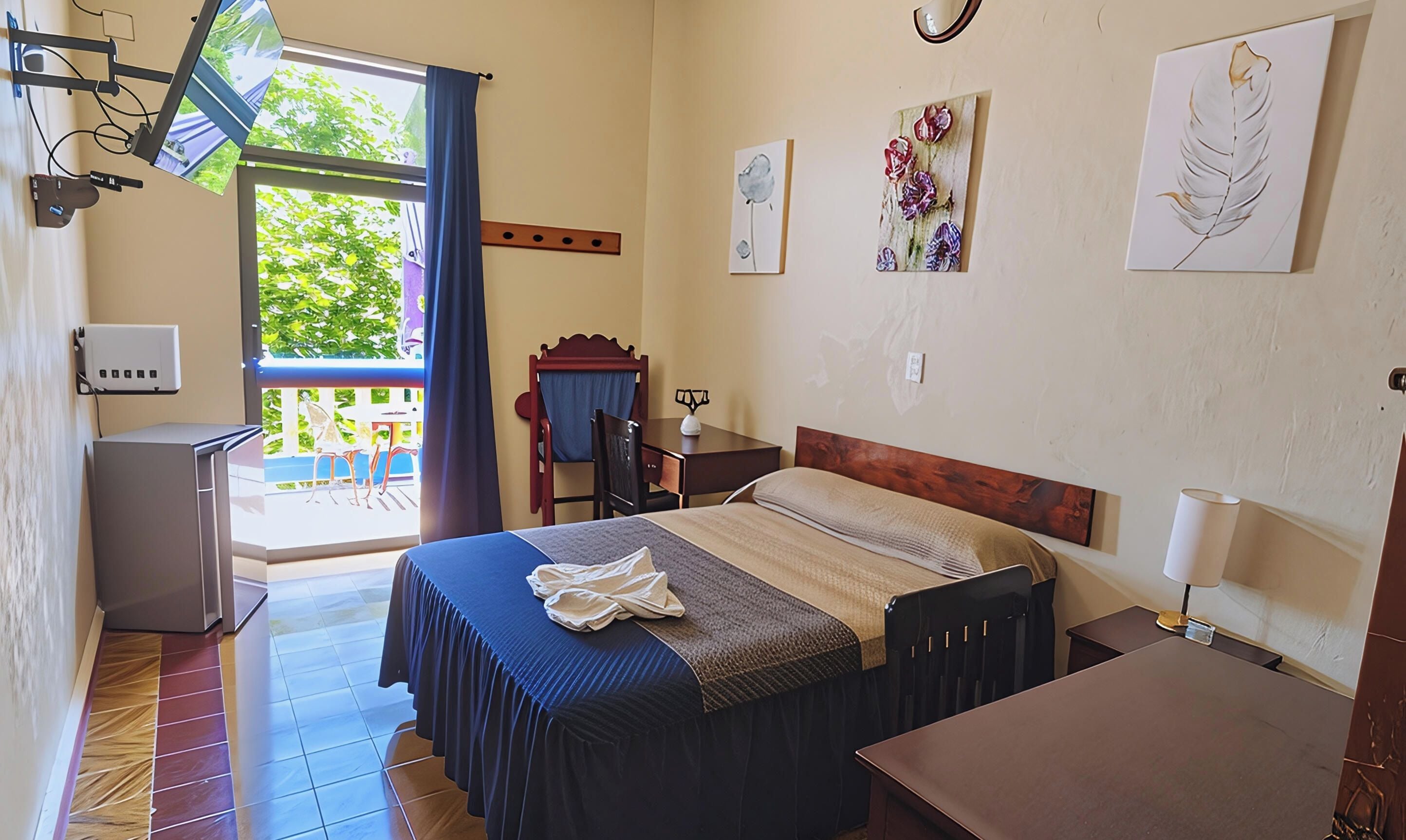 Habitación de hotel con cama, escritorio, silla, mesita, lámpara, obras de arte en las paredes, ventana con cortinas y balcón con mobiliario exterior.