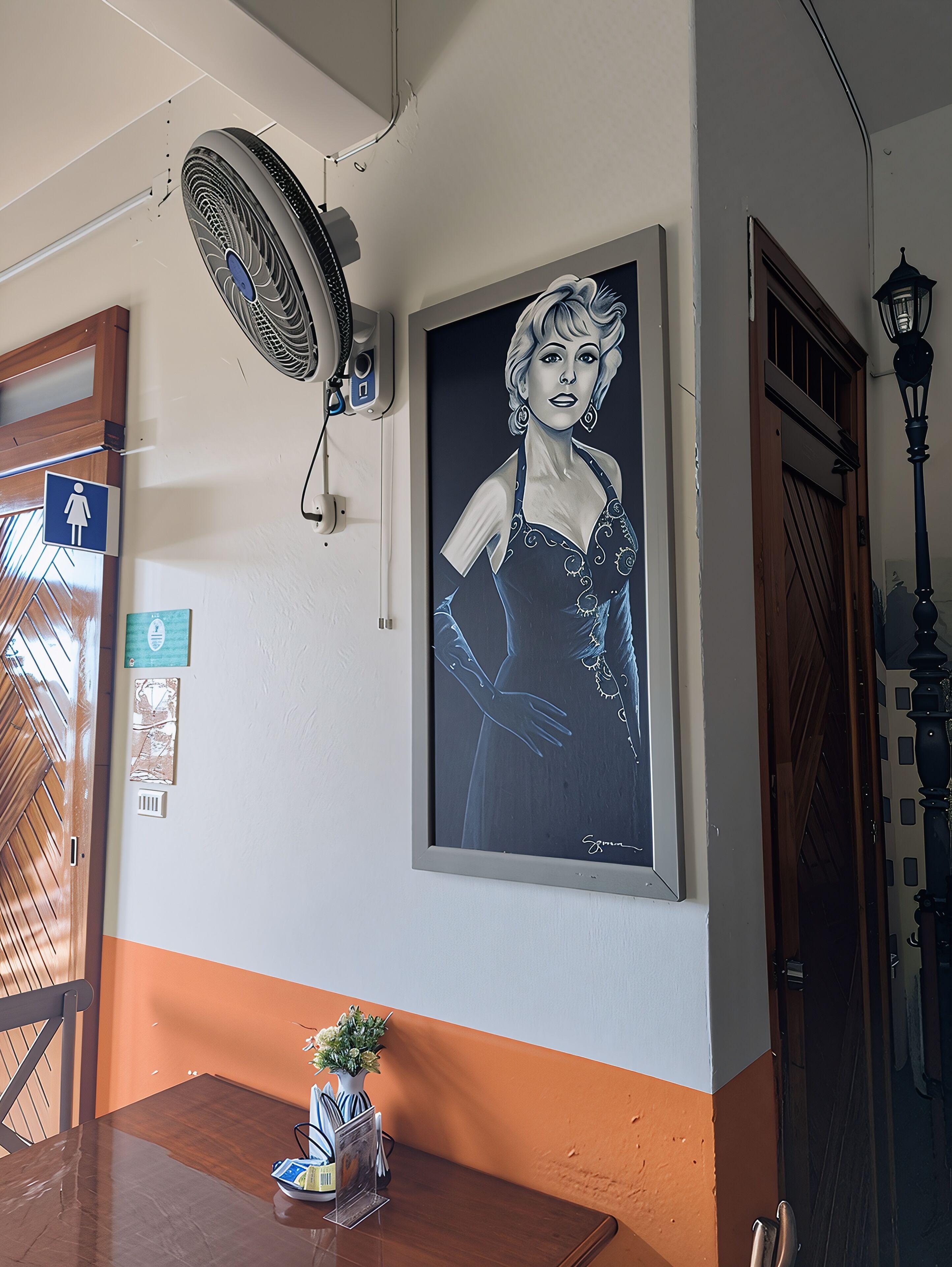 Paredes com um retrato emoldurado, um ventilador de teto, uma porta com olho mágico e uma pequena mesa com um vaso e revistas.