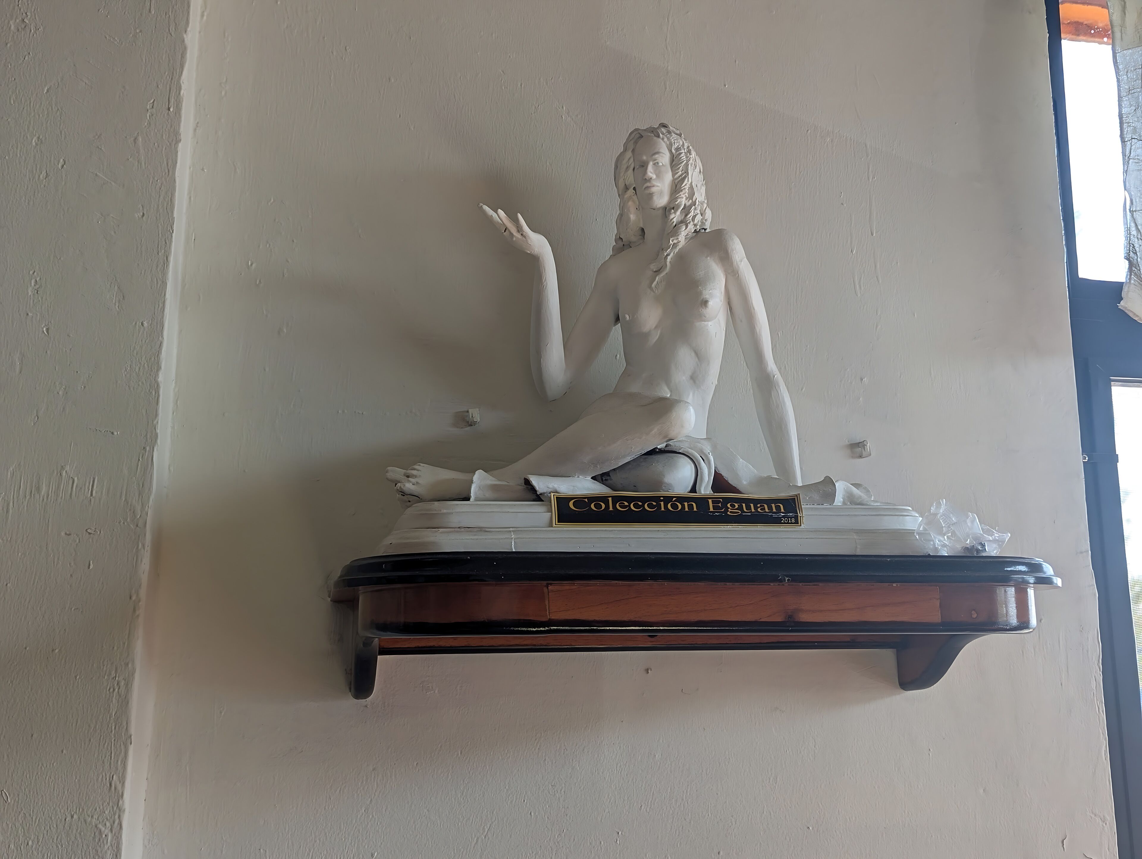 Escultura de una figura reclinada sobre una base de madera con una placa que dice 'Colección Eguan 2018'.