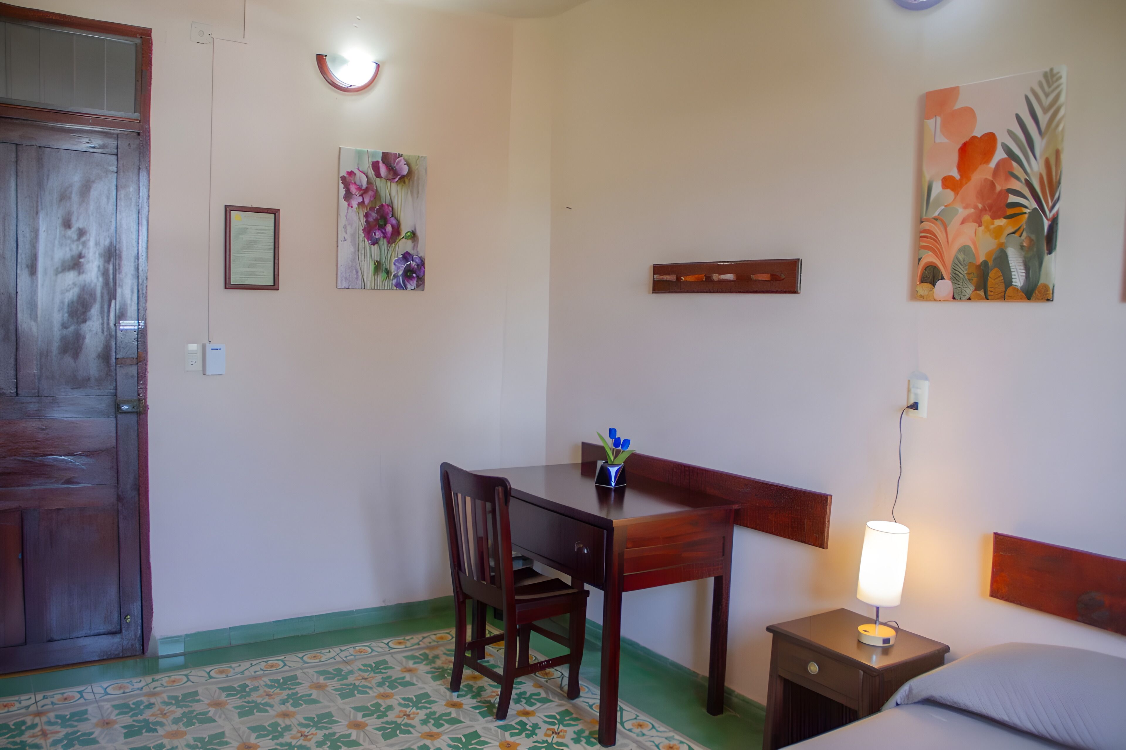 A room with a wooden desk, chair, bedside table with a lamp, and a bed.