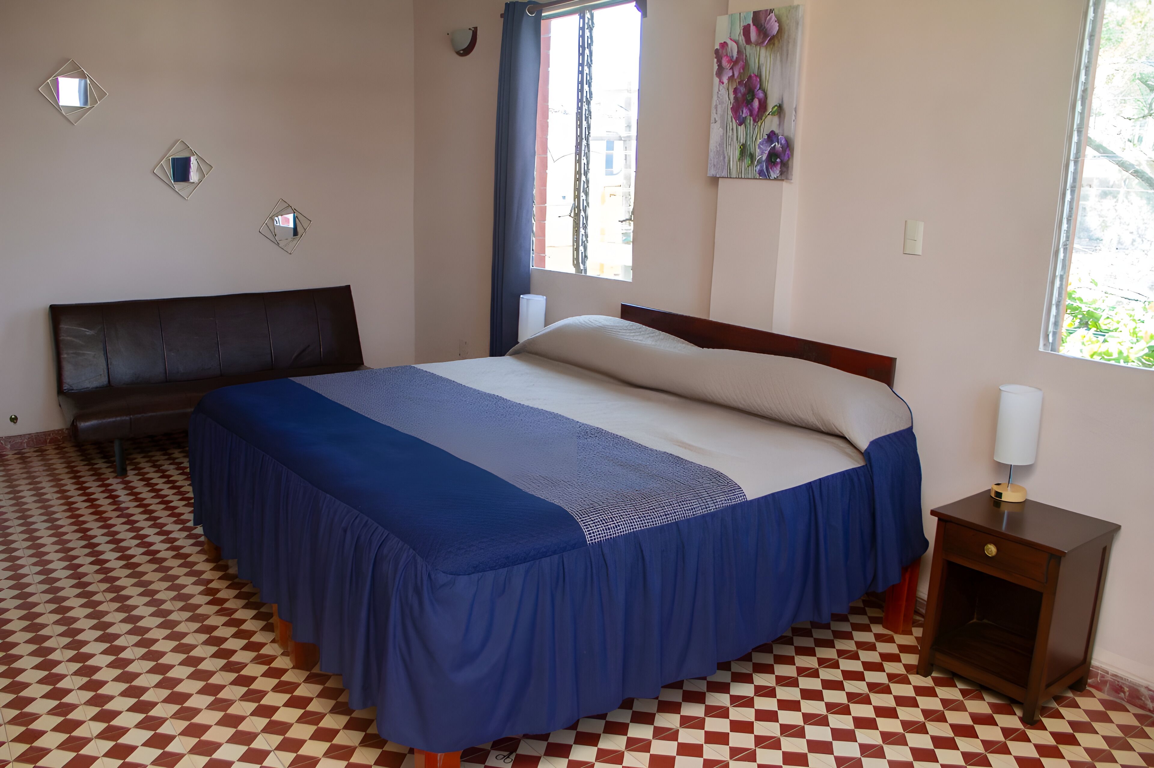 Un dormitorio con piso a cuadros, una cama con cubrecama azul, una mesita de noche con lámpara y una ventana con cortinas.