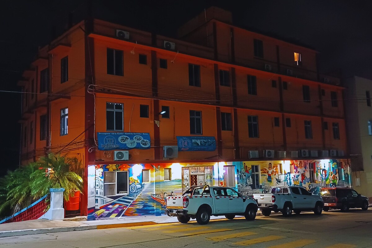 Un immeuble à plusieurs étages dont le rez-de-chaussée, bien éclairé, présente une fresque murale et une enseigne indiquant un restaurant. Deux véhicules sont garés devant.