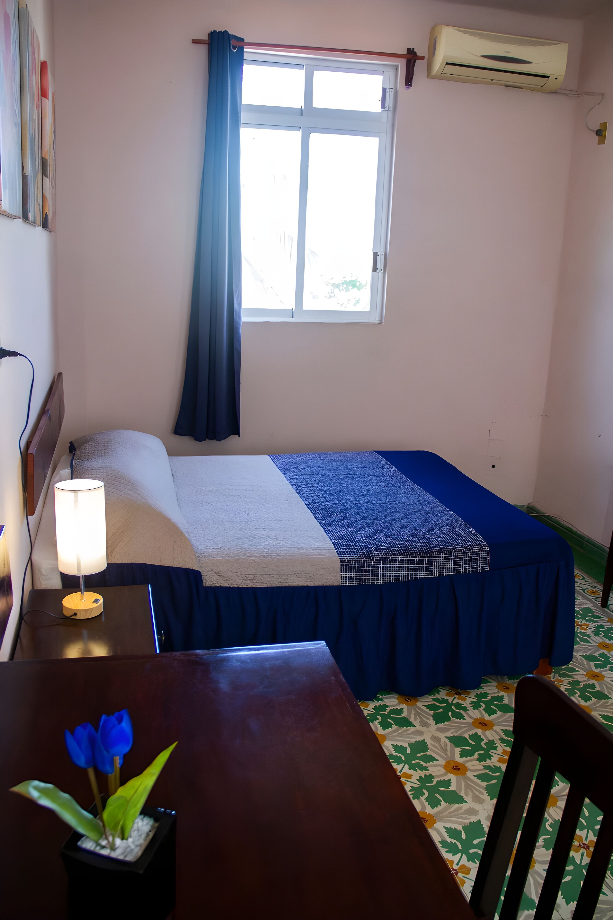 A small, neatly arranged bedroom with a single bed, a bedside table with a lamp, a chair, and a window with curtains.