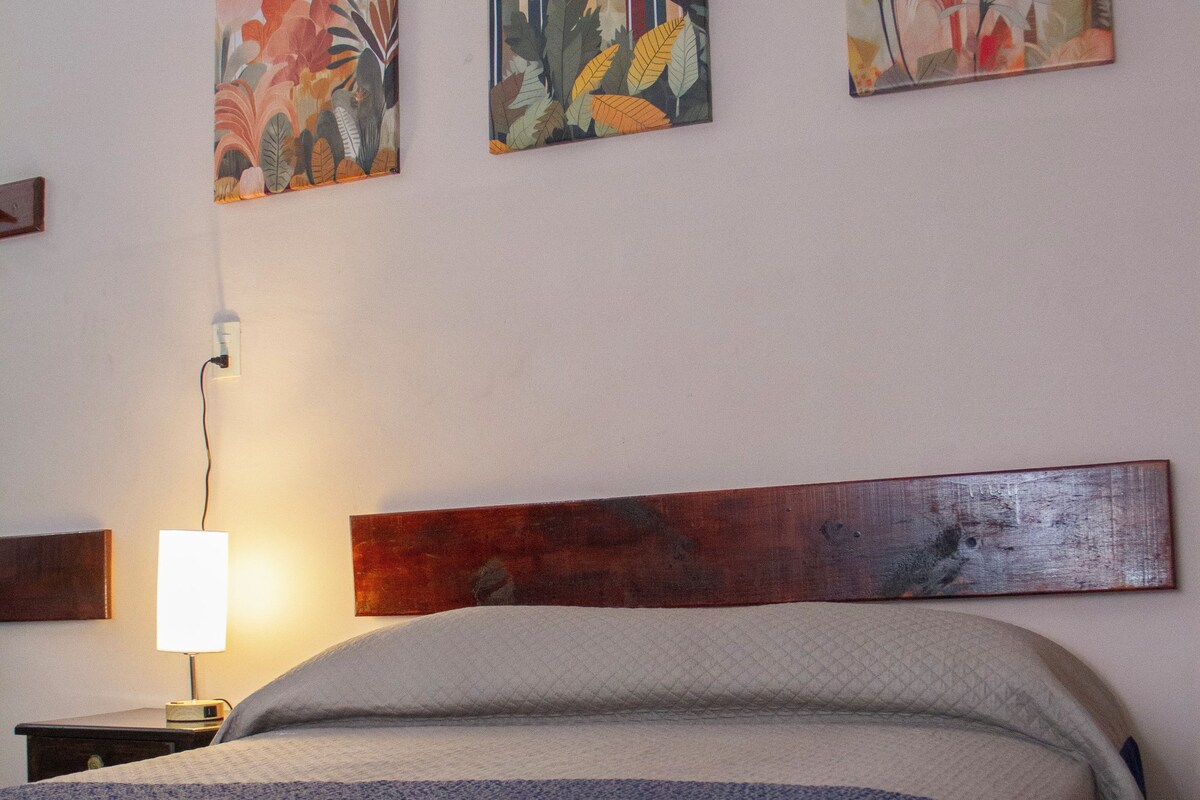 Quarto com uma cama, uma mesinha de cabeceira, uma luminária na parede e três quadros florais.