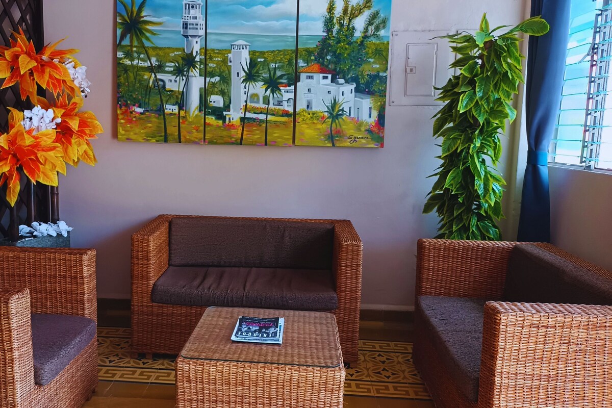 Une pièce meublée de mobilier en osier, ornée d’un tableau représentant un paysage côtier et agrémentée d’une plante en pot.