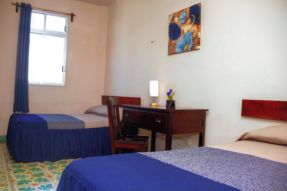 Een hotelkamer met twee bedden, een bureau, een stoel, een raam met gordijnen en een schilderij aan de muur.