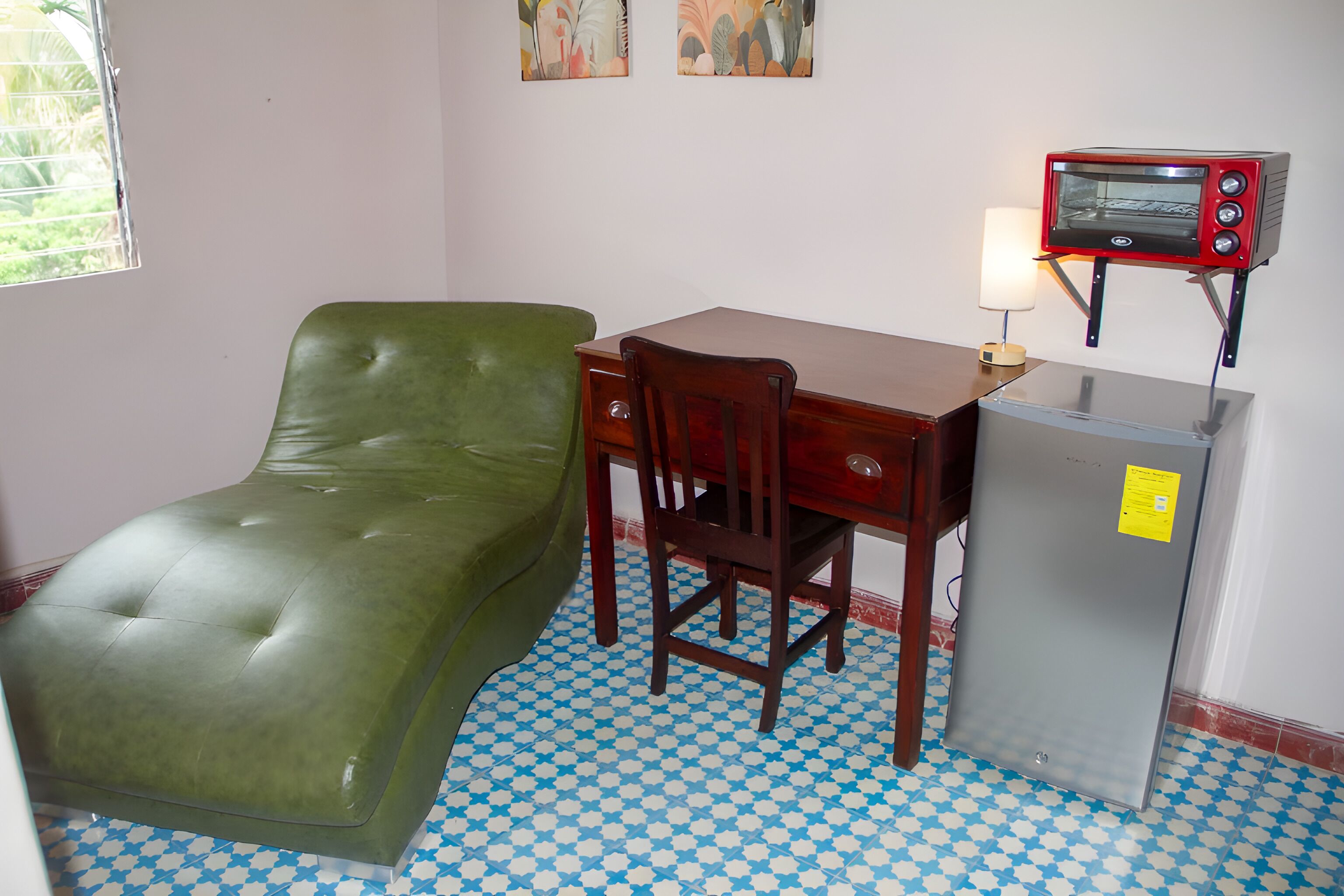A room with a green sofa, a wooden desk, a small refrigerator, and a wall-mounted television.