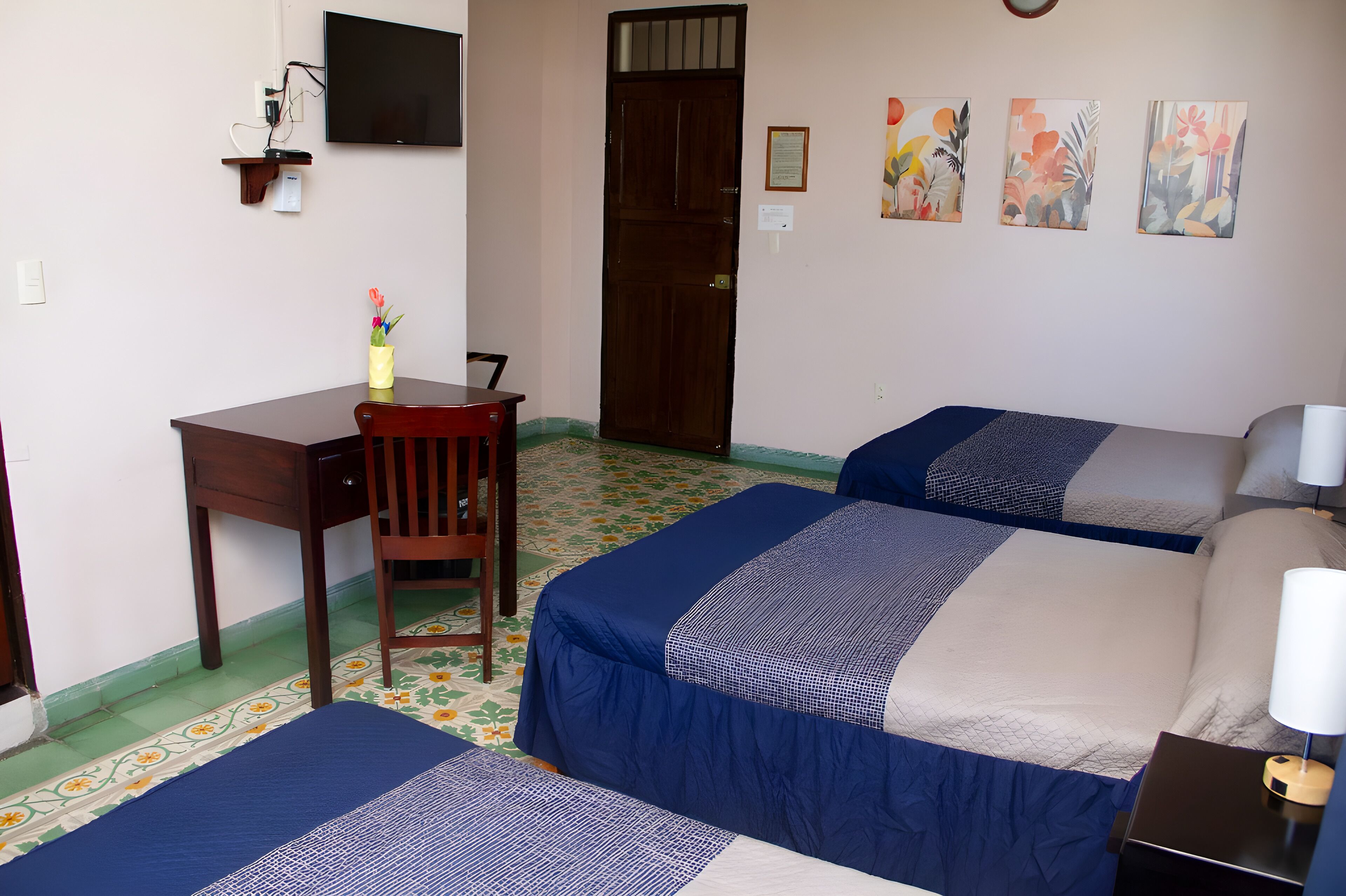 A hotel room with two beds, a desk, a chair, a TV, and floral wall art.