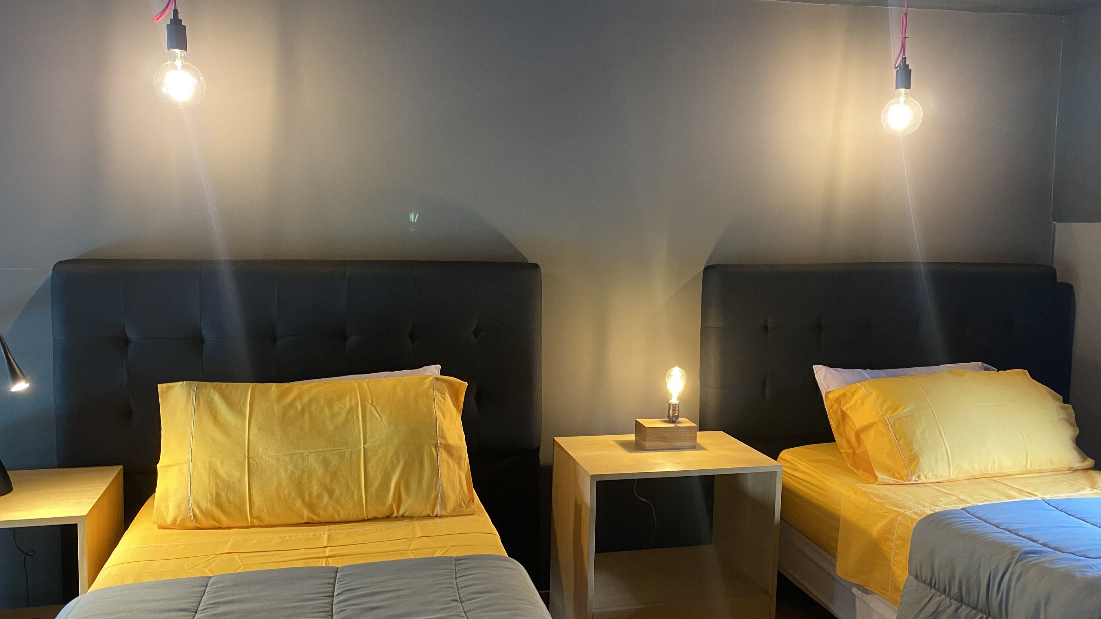 A hotel room with two beds, each with a headboard and a bedside table. There is a lamp on one of the tables.
