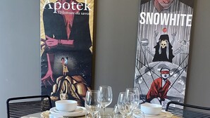 Une table de salle à manger ronde est dressée avec de l’assiette blanche, des bols et des verres à vin, et entourée de chaises noires agrémentées de sets de table en fibres tressées. Deux grandes œuvres d’art sont exposées sur le mur derrière la table.