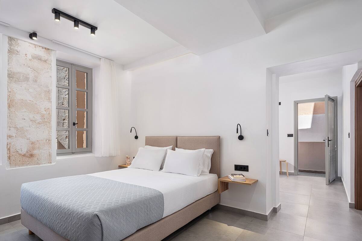 Un dormitorio moderno con una cama grande, mesitas de noche, una ventana con cortinas y un pasillo que lleva a otra habitación.
