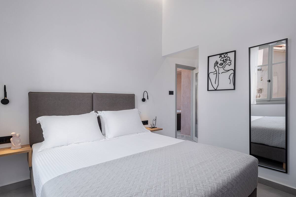 Una habitación de hotel moderna con una cama grande, una mesita de noche, un espejo y una puerta que da al baño.