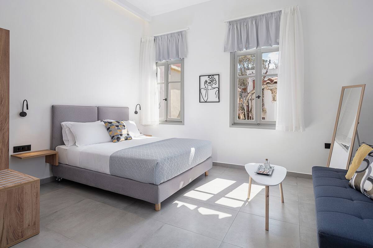 Un dormitorio moderno con cama, mesitas de noche, una pequeña mesa, un sofá y dos ventanas con cortinas.