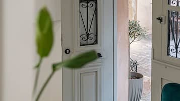 Une porte blanche avec un portail en métal, un pot de fleurs et une chaise de couleur turquoise dans un couloir au sol en marbre blanc.