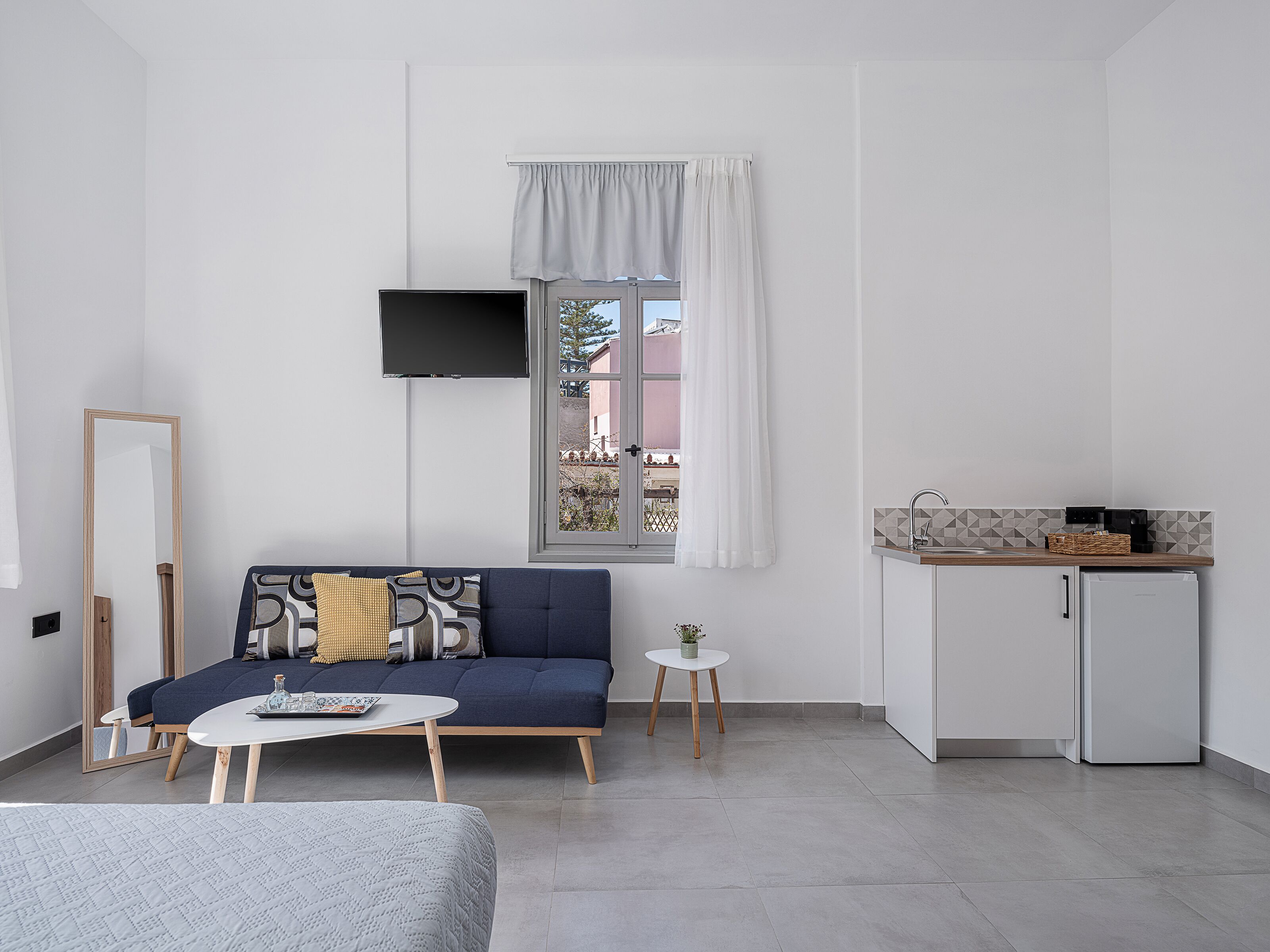 Ein modernes, minimalistisches Zimmer mit einer Couch, einem kleinen Couchtisch, einem an der Wand befestigten Fernseher, einem Fenster mit Vorhängen und einer Küchenzeile.