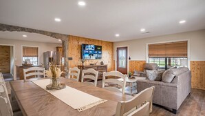 A spacious living area with a wooden dining table, white chairs, a grey sofa, and a TV mounted on the wall. - 3 Bedroom House w/ Outdoor Entertainment Area & Horse Accommodations (Bryan)