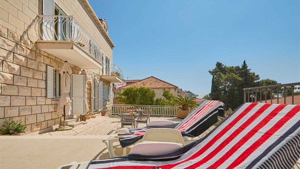 A sunlit outdoor seating area with lounge chairs and a table, a stone building with shutters, and a clear blue sky.