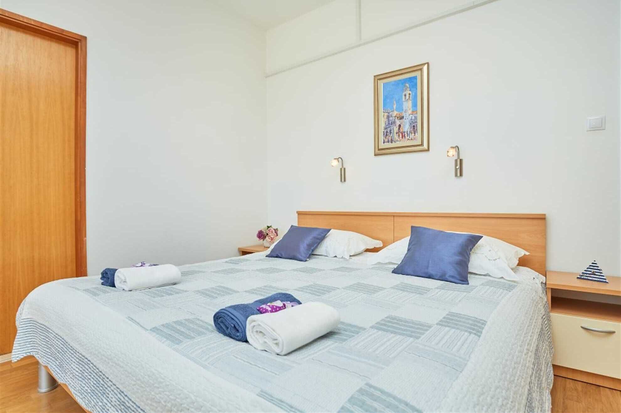 A neatly made bed with a quilt, pillows, and rolled towels. There is a nightstand with a lamp and a framed picture on the wall.