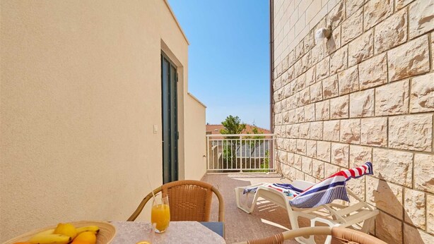 Un balcon avec des fauteuils en osier, une table sur laquelle se trouve un panier de fruits et un verre de jus d’orange, et un mur en pierre.