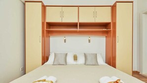 Un lit bien fait, avec deux serviettes enroulées, une tête de lit en bois agrémentée de deux placards et un mur de couleur claire.