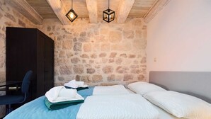 A bedroom with a stone wall, a bed with white linens, a desk with a chair, and two hanging lights. - Dominium Premium Location Suites Duplex apartment 1 (Dubrovnik)