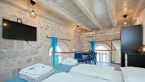 Un dormitorio con paredes de piedra, una cama con ropa blanca, un escritorio con sillas y un televisor montado en la pared.