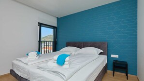 A neatly made bed with white linens and a blue towel, a small bedside table, and a window with a view of a building and a hill.