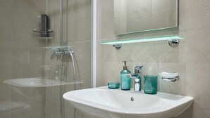 A bathroom with a glass shower enclosure, a white sink with a glass shelf above it, and a mirror.