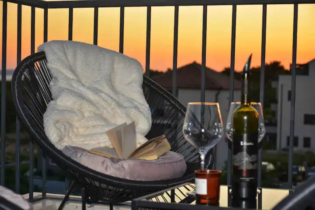 En balkon med en fletstol, pude, bog, to vinglas og en flaske vin.